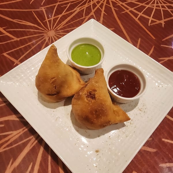 Two golden-brown samosas on a white rectangular plate with green and red chutneys.