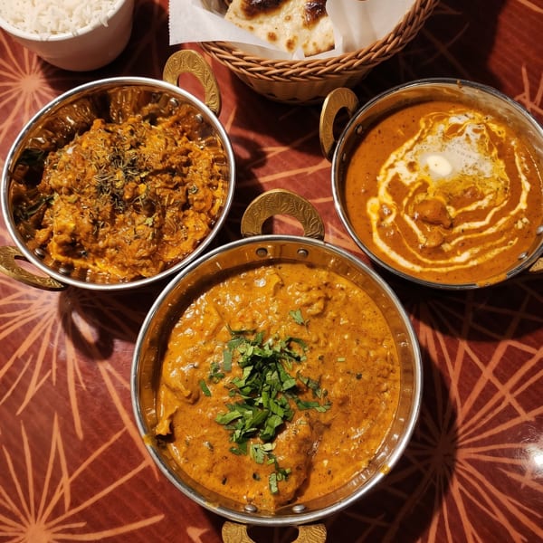 Three bowls of Indian curry, a basket of naan bread, and a cup of rice on a patterned table.