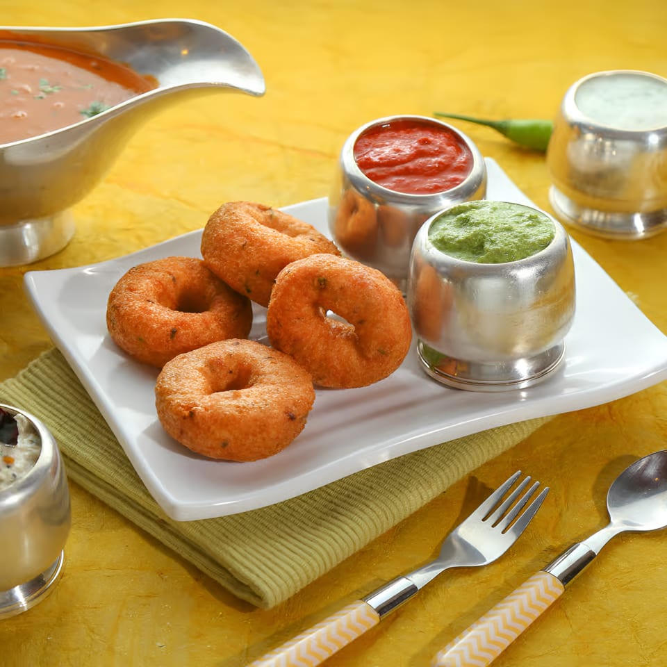 Golden-brown vada donuts on a white plate with various chutneys and sambar in silver bowls.