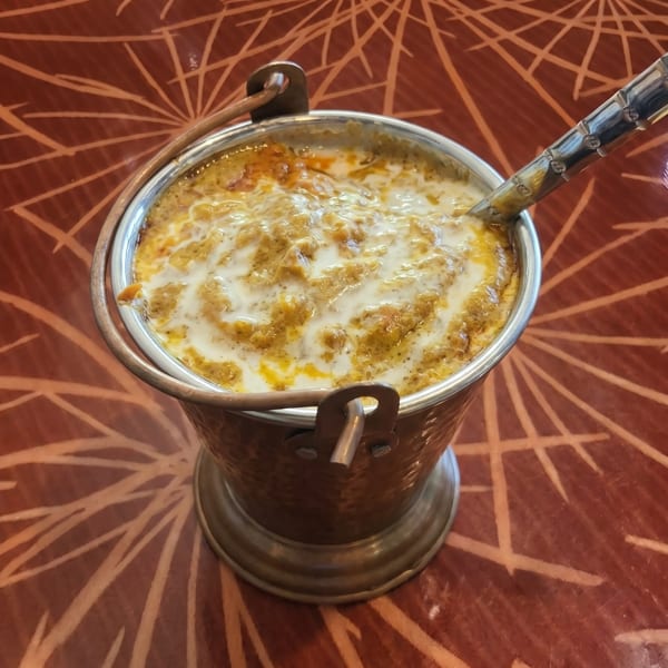 A copper bucket filled with creamy Indian curry, drizzled with white sauce, on a patterned red table.