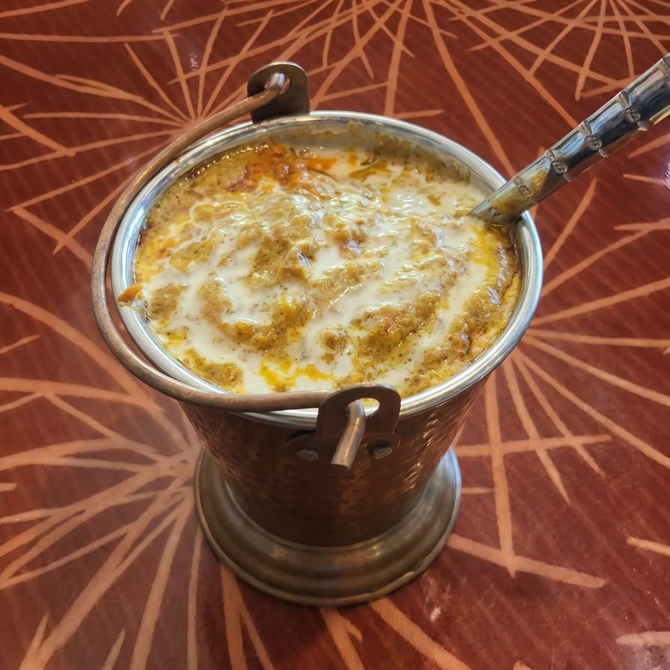 A copper bucket filled with creamy Indian curry, drizzled with white sauce, on a patterned red table.