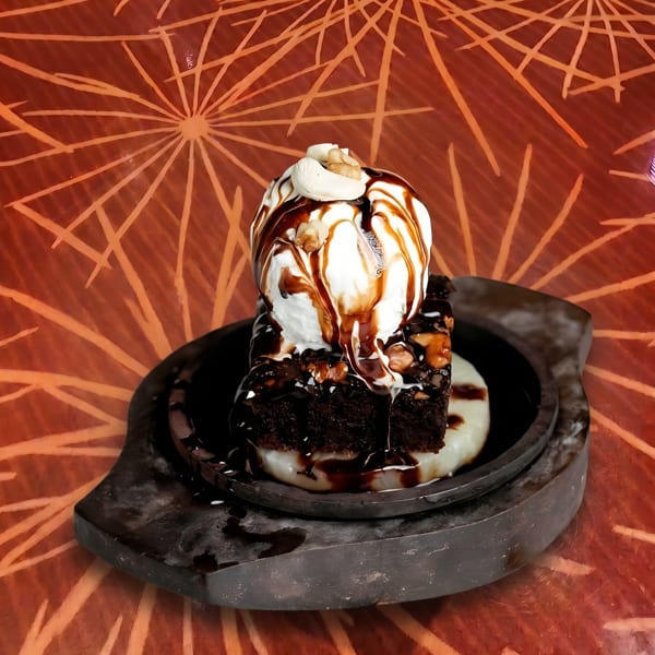 A sizzling brownie with vanilla ice cream, chocolate sauce, and nuts on a dark plate.