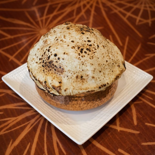 A golden pot of biryani, sealed with a charred flatbread, sits on a white plate.