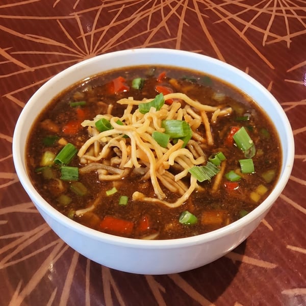 A white bowl of dark soup with noodles, red peppers, and green onions on a patterned table.